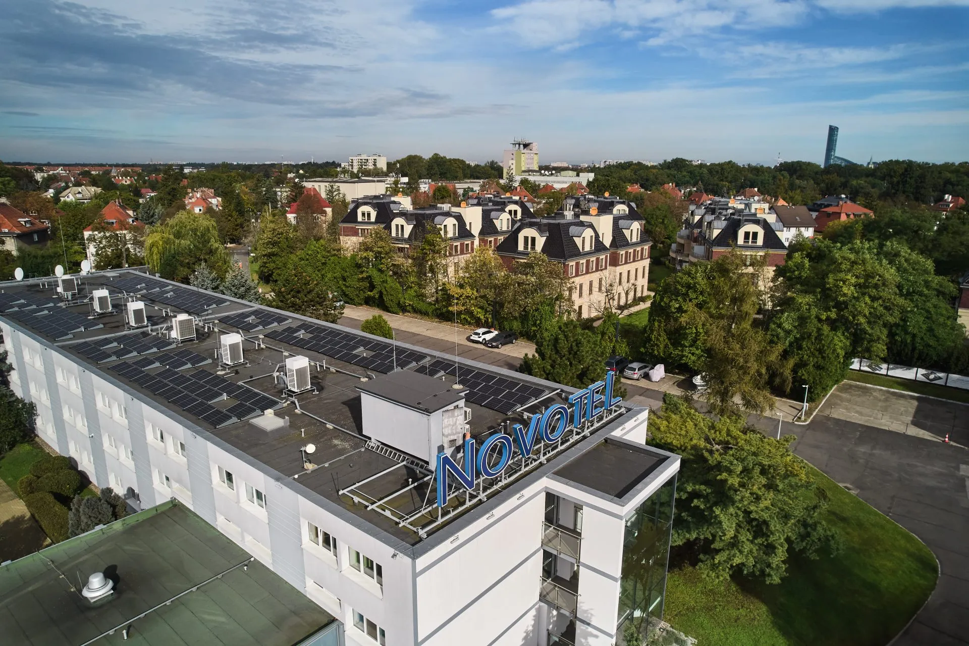 Widok z góry na budynek oznaczony niebieskim napisem „Novotel”, umieszczonym na dachu. W tle rozciągają się drzewa, osiedla i samotna wieża. Niebo jest lekko zachmurzone, nadając scenie spokojny nastrój. Hotel znajduje się pośrodku zielonego, miejskiego krajobrazu