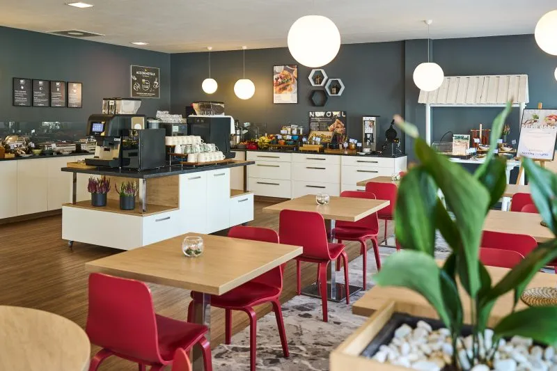 Wnętrze eleganckiej restauracji z czerwonymi krzesłami i drewnianymi stołami. Na ścianach wiszą obrazy, a sufit ozdabiają okrągłe lampy. W głębi znajduje się bar z napojami. Atmosfera jest ciepła i przytulna.