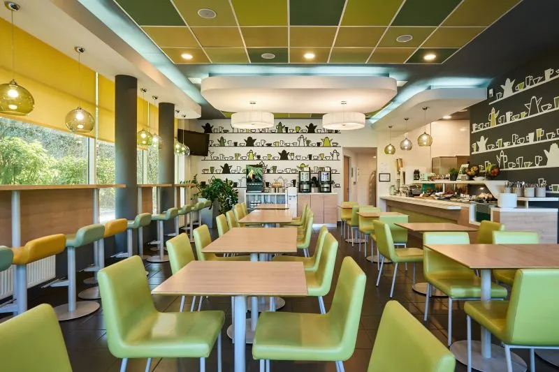 Zdjęcie przedstawia przestronną i nowoczesną salę restauracyjną w hotelu. Pomieszczenie urządzone jest w jasnych barwach, z dominującą zielenią i drewnem. Stoły i krzesła ustawione są w równych rzędach, a na ścianach zawieszono grafiki i półki z filiżankami, co nadaje wnętrzu domowego klimatu. Duże okna po lewej stronie wpuszczają dużo naturalnego światła, co sprawia, że przestrzeń jest jasna i przyjemna.