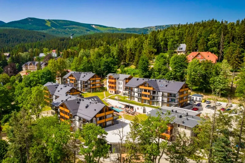 kompleks apartamentowy położony wśród zieleni u podnóża gór, otoczony lasem i górami w tle. Budynki mają drewniane akcenty elewacyjne, a w centralnej części znajduje się basen z leżakami.