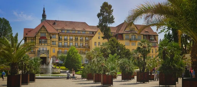 Imponujący budynek Domu Zdrojowego w Kudowie-Zdroju, otoczony egzotyczną roślinnością i fontanną na tle słonecznego nieba. Architektura nawiązuje do stylu tyrolskiego, tworząc wyjątkową atmosferę kurortu.