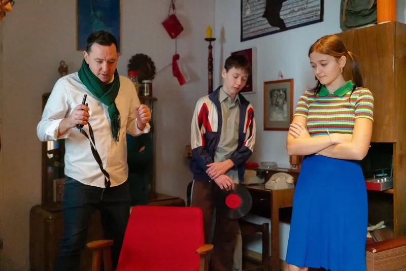 Mężczyzna z narzędziami rozmawia z dwojgiem młodych ludzi w mieszkaniu z minionej epoki, odtwarzając scenę do filmu Centrum Historii Zajezdnia.