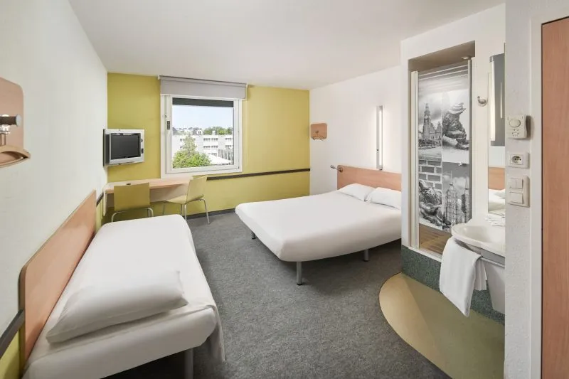 Obraz pokazuje typowy pokój hotelowy typu budget, urządzony minimalistycznie, ale funkcjonalnie. W pokoju znajdują się dwa pojedyncze łóżka ustawione prostopadle do siebie oraz niewielki blat z telewizorem na ścianie. Przy oknie widoczna jest zielona zasłona oraz kaloryfer. Z prawej strony zdjęcia znajduje się otwarte wejście do łazienki. Pokój jest jasny i schludny, utrzymany w białych i pastelowych kolorach.