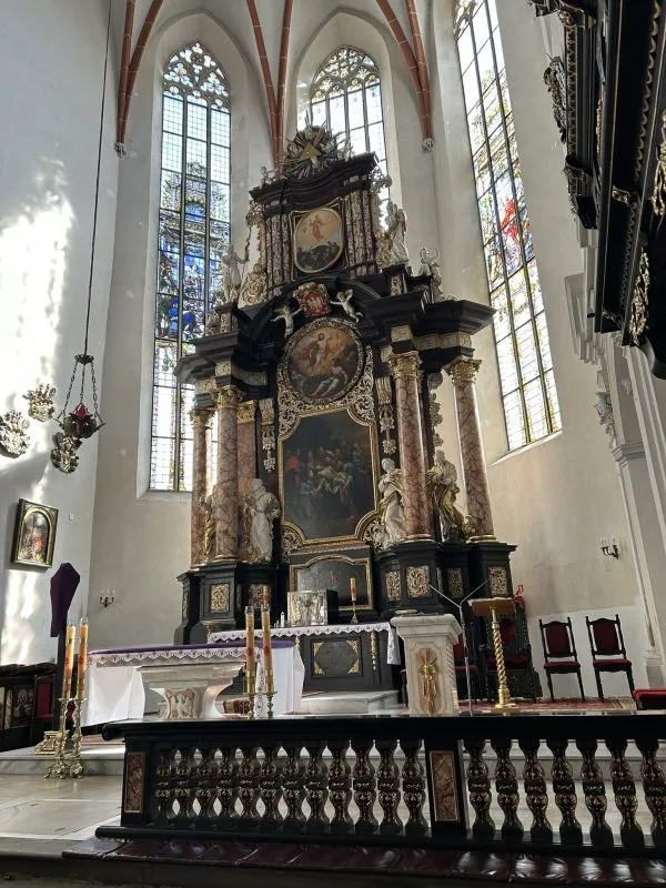 Wnętrze kościoła z barokowym ołtarzem głównym, ozdobionym złoceniami, rzeźbami i obrazami religijnymi. W tle wysokie, gotyckie okna z kolorowymi witrażami. Przed ołtarzem stoją świeczniki i ambona.