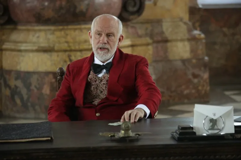 John Malkovich as an elegant older man in a red blazer sitting behind a heavy desk in an ornate interior from the movie "Valley of the Gods".