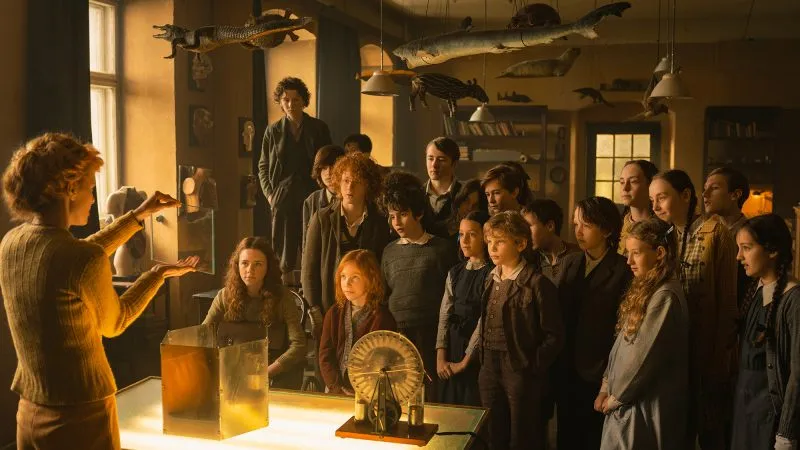 A still from the film "Kajtek Czarodziej" shows a teacher demonstrating an experiment at a table, surrounded by a focused group of students in an old classroom.