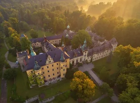 Rozległy Zamek Kliczków z wieżami, dziedzińcem i czerwonymi dachami widziany jest z lotu ptaka wśród gęstych drzew oświetlonych ciepłym słońcem.