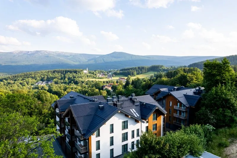 Malownicza panorama Szklarskiej Poręby z widokiem na pasmo Karkonoszy oraz kompleks budynków Happy Valley Resort wkomponowany w zielone otoczenie. W tle widać Śnieżne Kotły oraz charakterystyczną górę Szrenicę, co podkreśla górski, wypoczynkowy charakter miejsca.