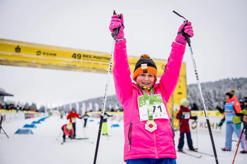 A child in a pink snowsuit raises ski poles above their head after finishing the Bieg Piastów, standing on the snow with a medal and a race bib.