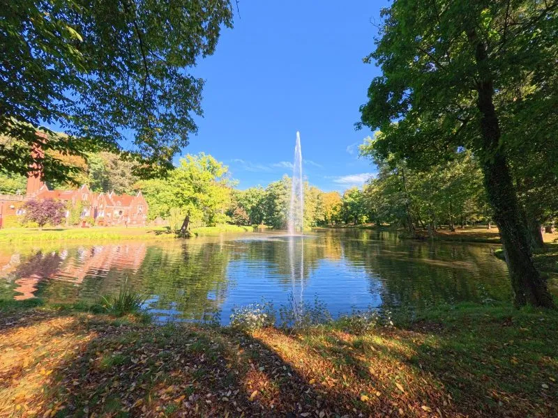 El estanque del parque en Kamieniec Ząbkowicki refleja la fuente, los árboles y una parte del palacio, y las orillas sombreadas están cubiertas de césped y hojas de otoño.
