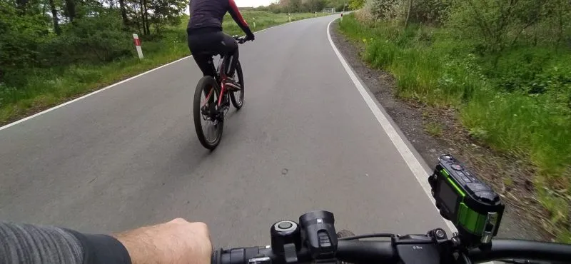 A cyclist seen from the perspective of another rider travels along the asphalt Ścinawa-Prochowice path, winding through green meadows, bushes, and fields.