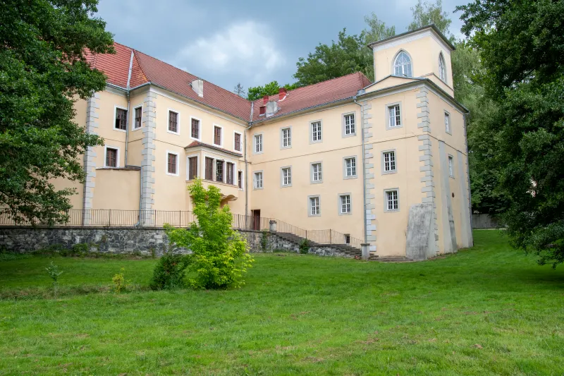 Na zdjęciu widoczny jest jasny, historyczny budynek o czerwonym dachu, otoczony zielenią i rozległym trawnikiem. Obiekt znajduje się w spokojnym, parkowym otoczeniu, a nad nim widać ciężkie, burzowe chmury.