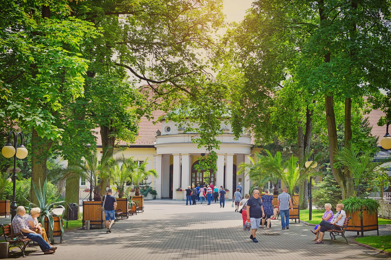 Szeroka aleja uzdrowiskowa prowadzi przez zielony park ku jasnemu pawilonowi, mijając ławki, spacerowiczów, stare drzewa i ozdobne rośliny.