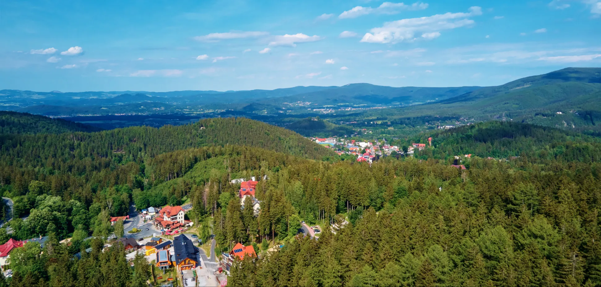 Panoramiczny widok z góry na miejscowość ukrytą w rozległych lasach, z czerwonymi dachami zabudowań i pasmami łagodnych gór na horyzoncie.