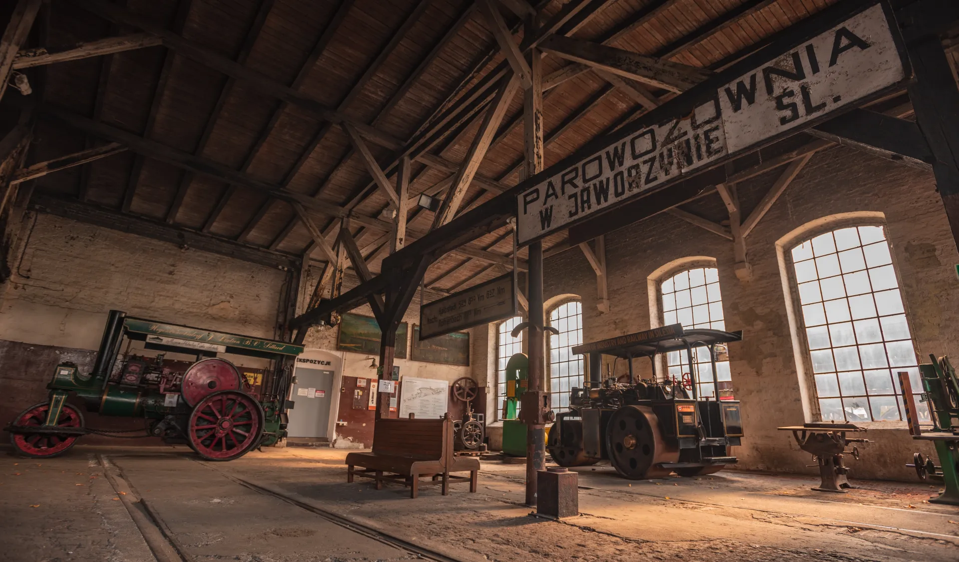 Zabytkowa hala Muzeum Kolejnictwa w Jaworzynie Śląskiej mieści historyczne maszyny parowe, warsztatowe urządzenia i drewniane ławy oświetlone przez wysokie okna.