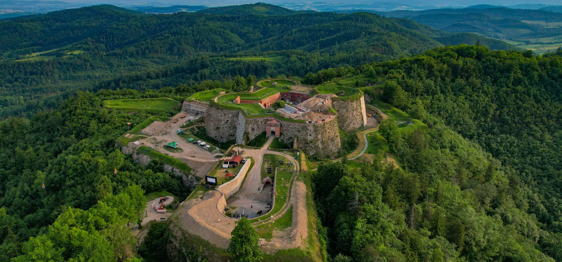 Die Vogelperspektive auf die Festung Silberberg zeigt mächtige steinere Bastionen und Innenhöfe, umgeben von grünen Hügeln und einer Berglandschaft.