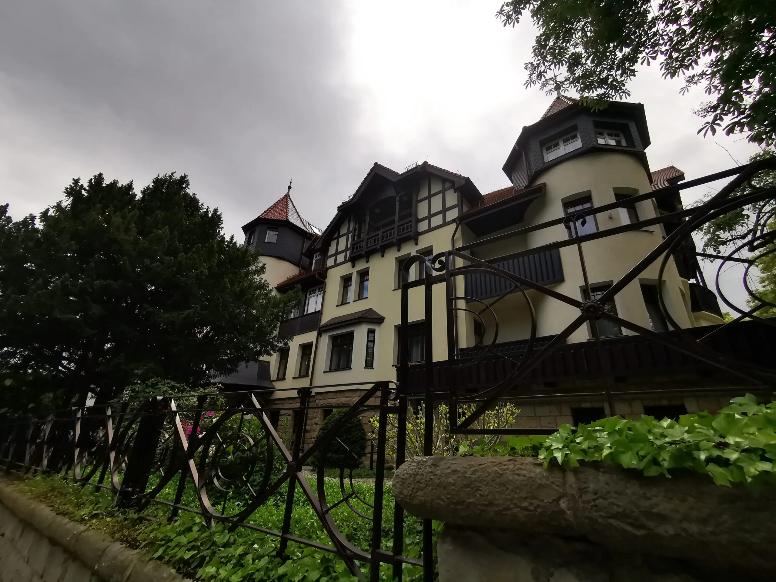 Hotel Bukowy Park v Polanici-Zdroji má světlou fasádu s tmavými dřevěnými detaily, balkony a věžičkami, obklopenou mokrou zelení zahrady.