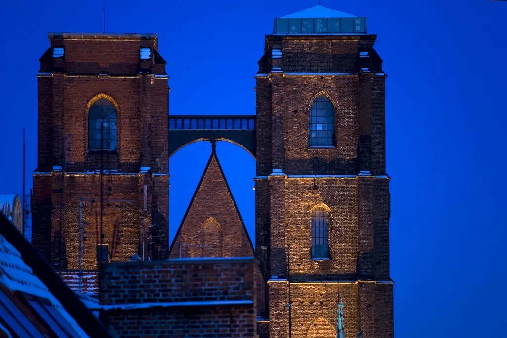 Widok na murowane, gotyckie wieże z widocznym mostkiem łączącym je na znacznej wysokości na tle wieczornego nieba