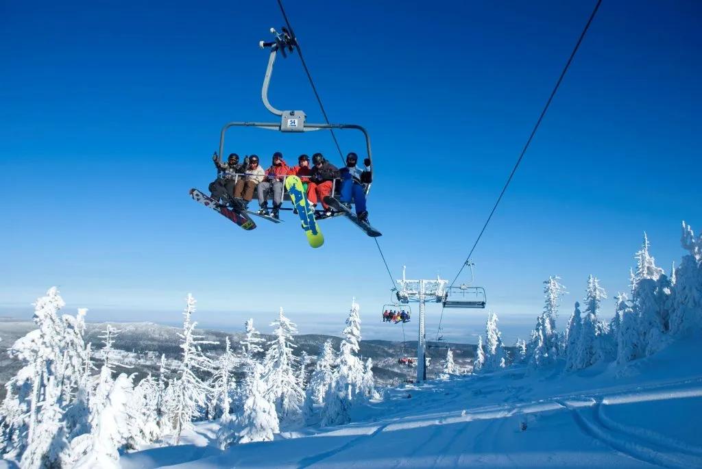 Panorama górska z kolejką linową na pierwszym planie. Korzystają z niej narciarze i snowboardziści ubrani w kolorowe kombinezony. Drzewa są oblodzone, a stok ośnieżony.