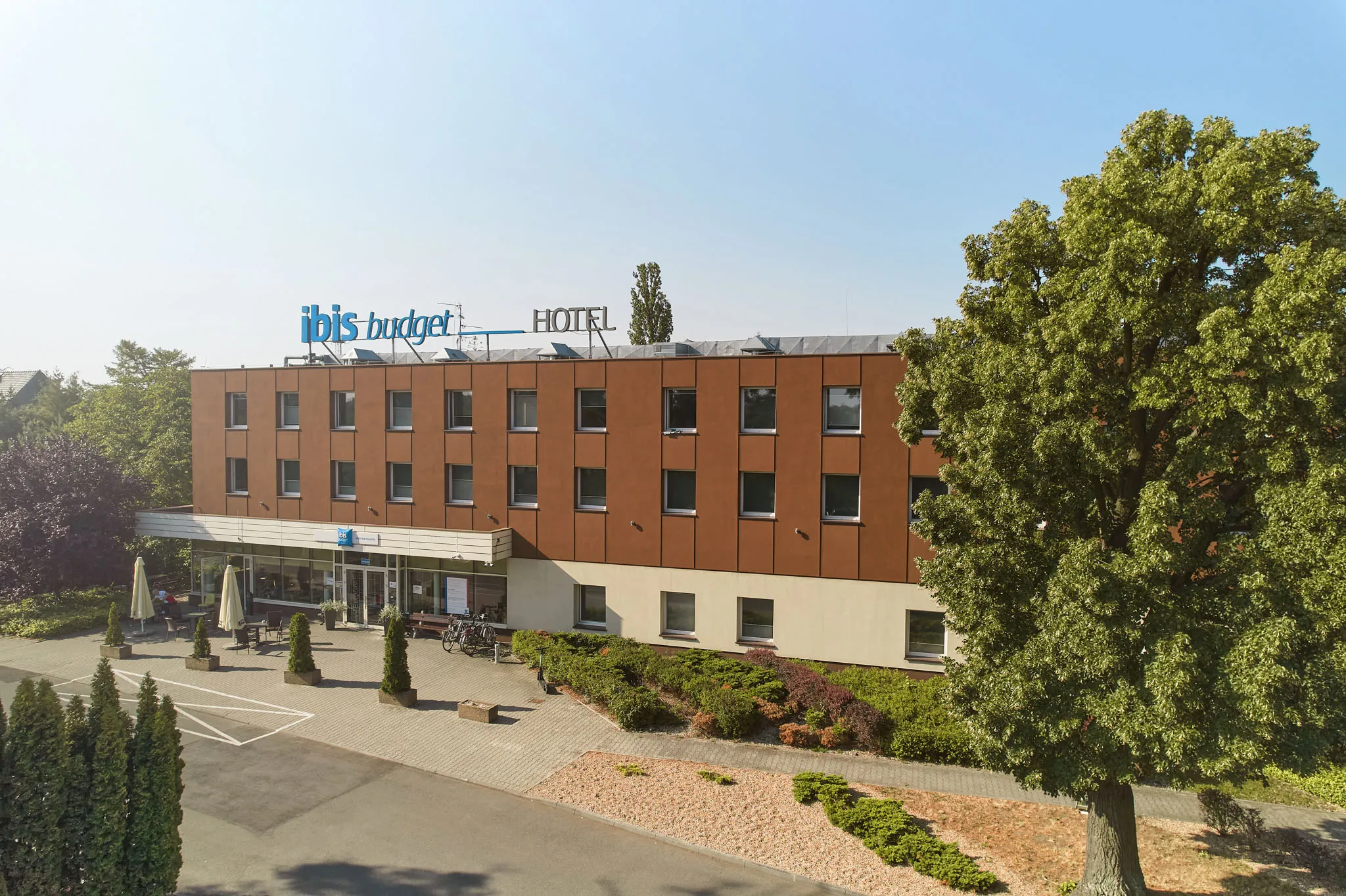 Na zdjęciu widać zewnętrzną fasadę hotelu ibis budget, zbudowaną z czerwonej cegły. Budynek ma kilka pięter, a jego bryła jest prosta i nowoczesna. Przed wejściem znajdują się starannie przycięte krzewy oraz podjazd, na którym zaparkowano kilka samochodów. W tle rosną wysokie drzewa, co nadaje miejscu spokojny, zielony charakter.