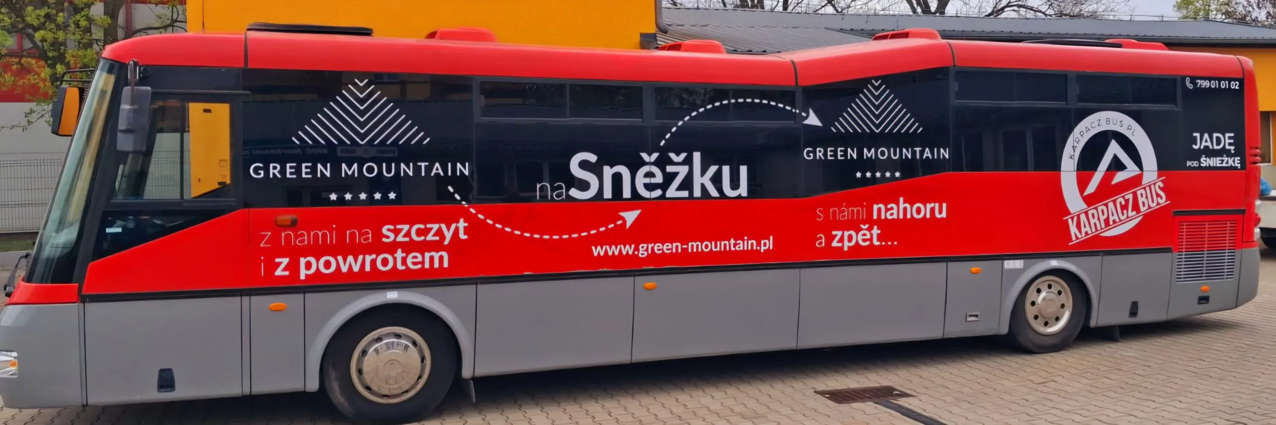 Zdjęcie przedstawia widok boczny na autobus turystyczny w kolorze czerwono-szarym